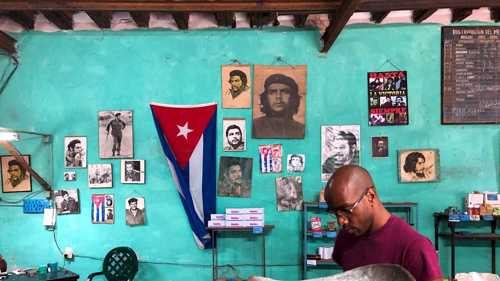 Image shows a young Cuban man with glasses sitting with what look to be large weighing scales. He is in front of a turquoise wall with a Cuban flag and images of Che Guevara and a young Fidel Castro.